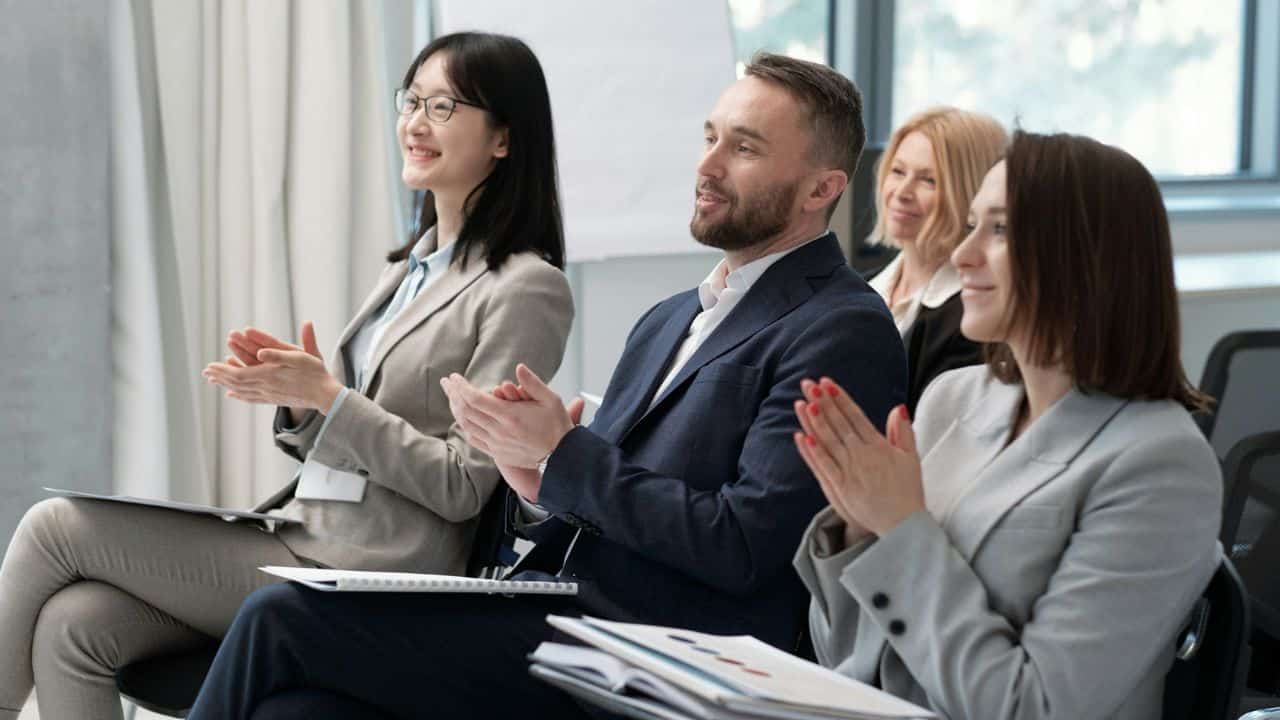 A diverse group of smiling professionals are clapping while seated at a conference.