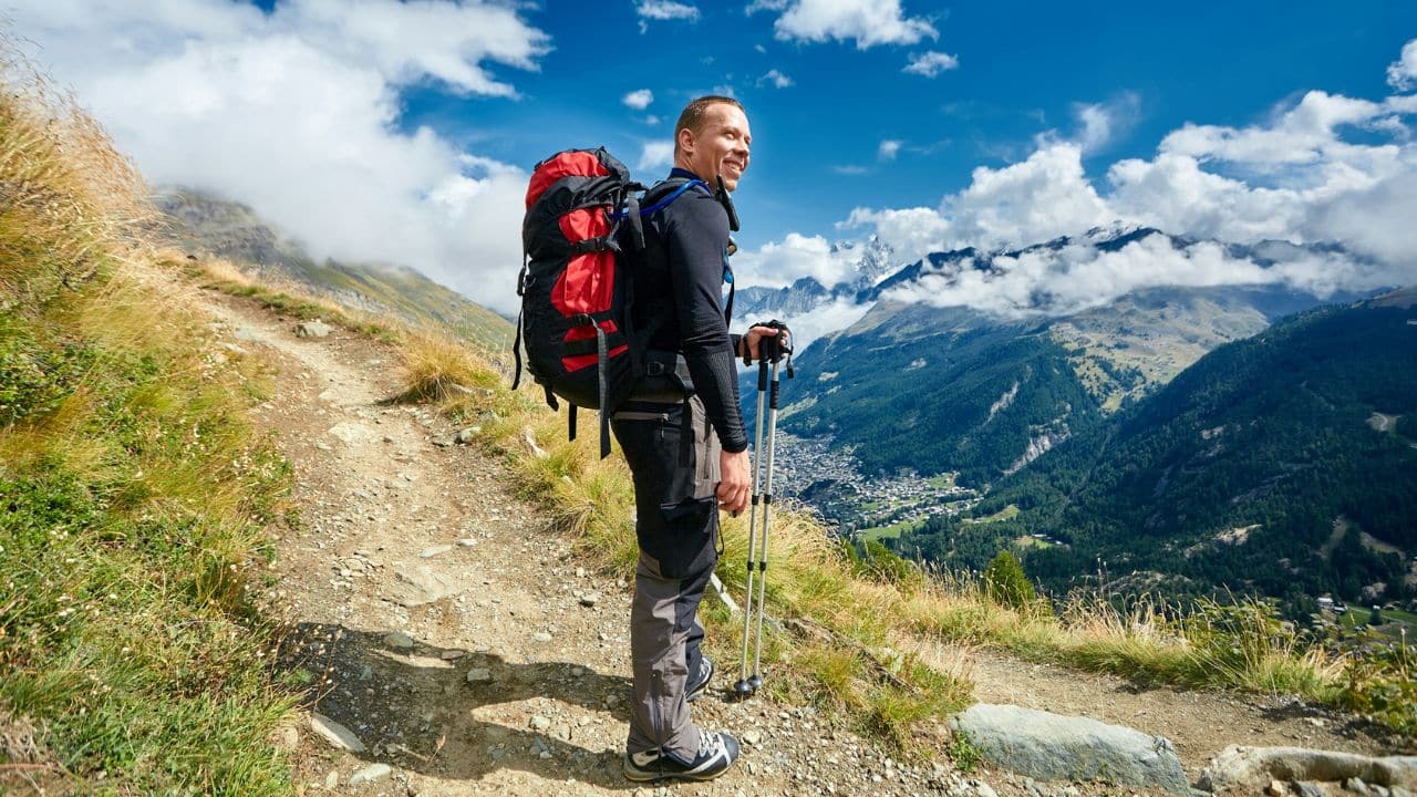 A smiling hiker with a large backpack stands on a mountain trail, looking away.