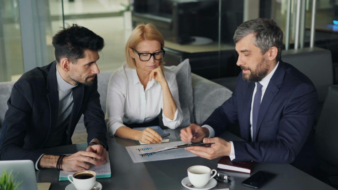 Three serious-looking business professionals review a financial report together at a table.