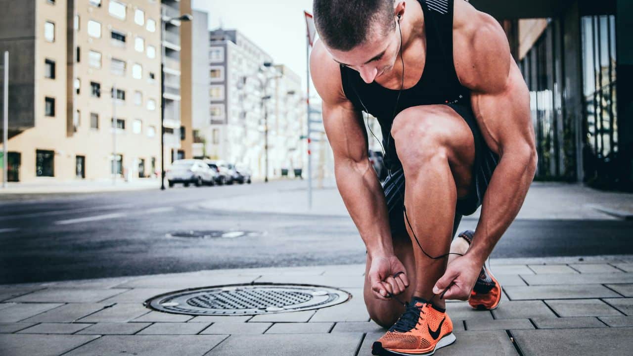 A fit, muscular man wearing headphones ties his sneaker on a city sidewalk.