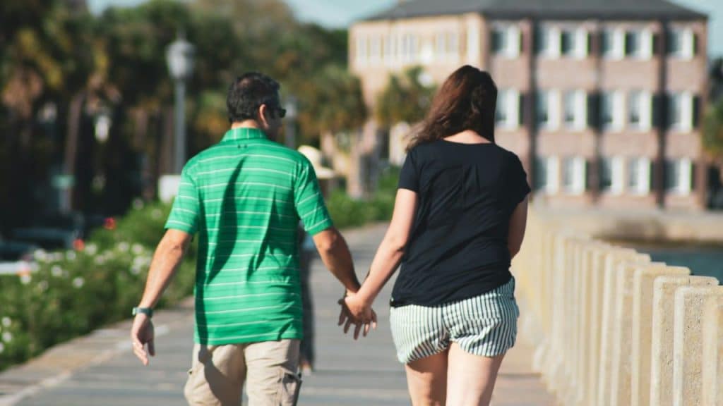 A middle-aged couple holds hands and walks down a path on a sunny day.