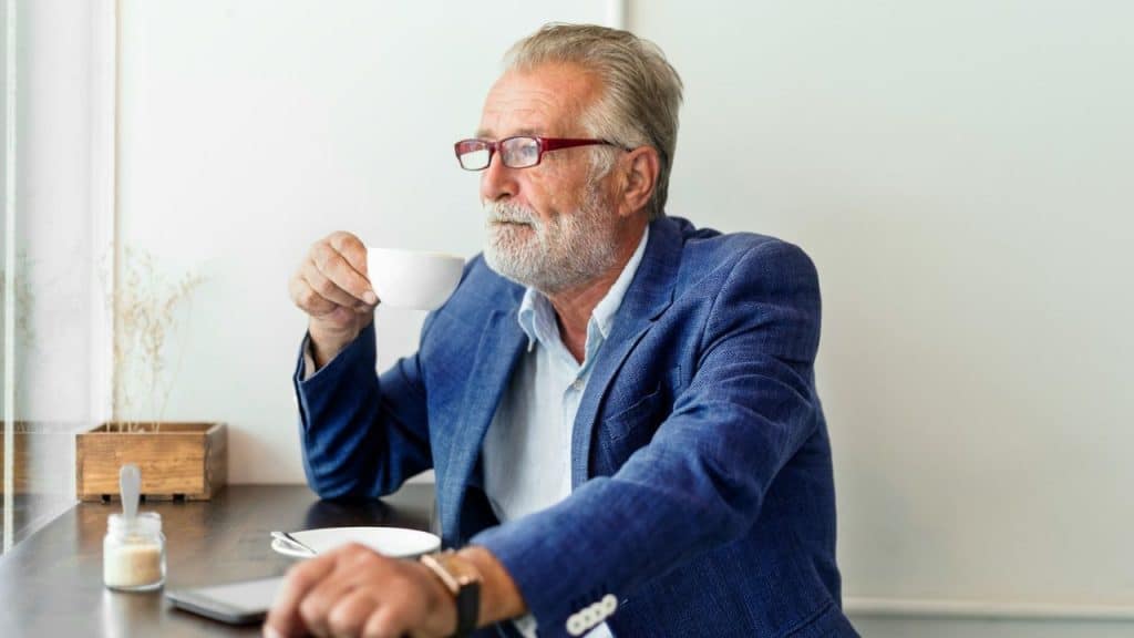 A contemplative, older man with a beard and glasses sips coffee while sitting at a table.