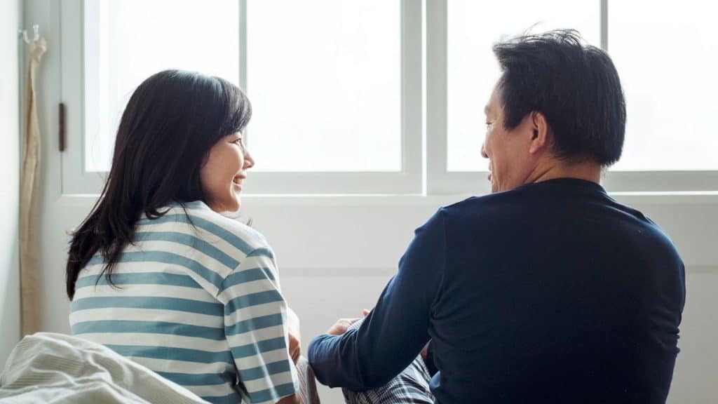 A smiling couple, seen from behind, sits on a bed and talks to each other.