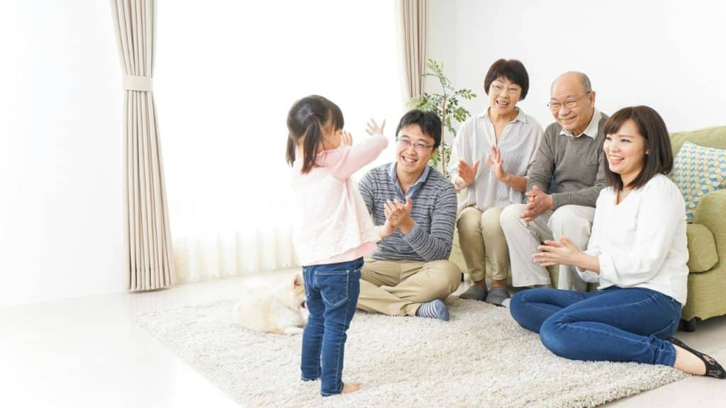A happy, multigenerational family watches and claps for a young girl playing in a living room.