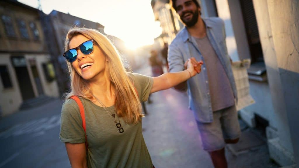 A happy blonde woman in sunglasses leads a man by the hand on a sunny street.