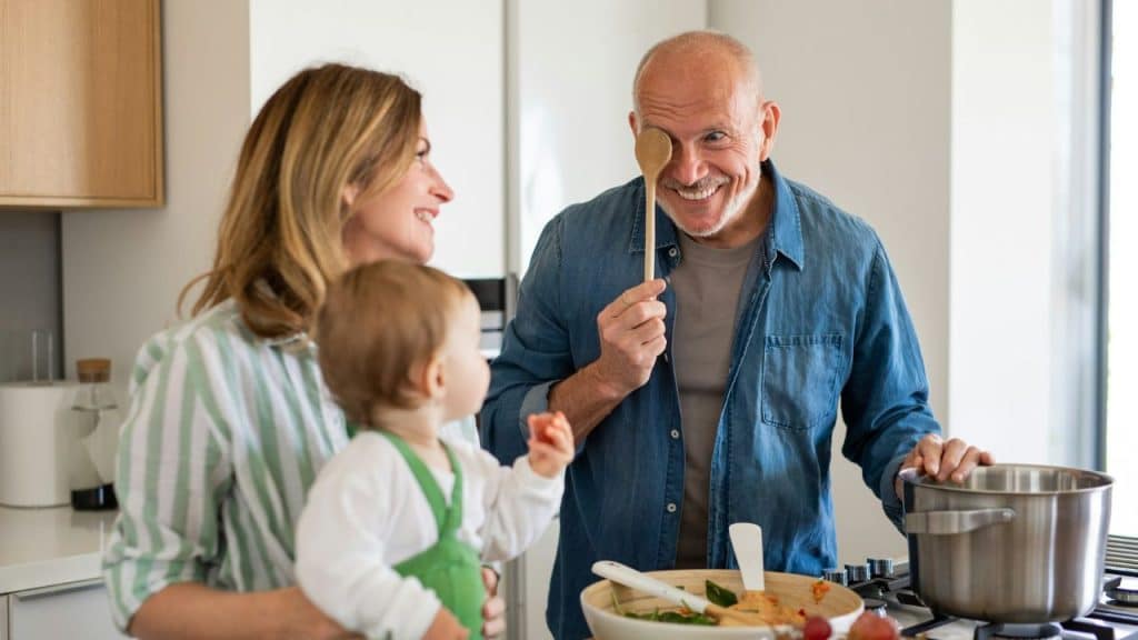 A joyful family is cooking in a kitchen, with a grandfather making a funny face at a baby.