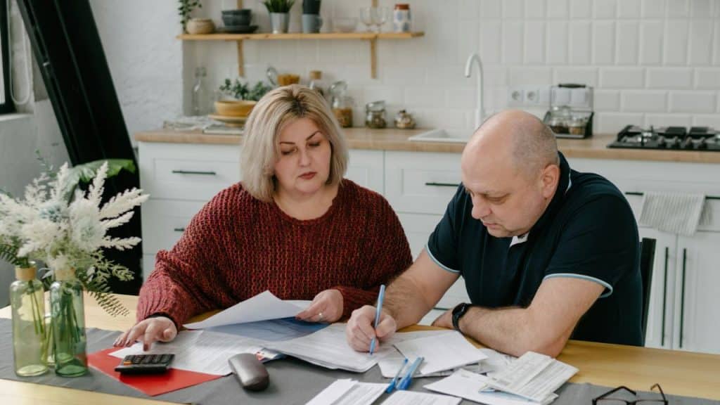 A serious middle-aged couple is sitting at a kitchen table, looking at financial documents.