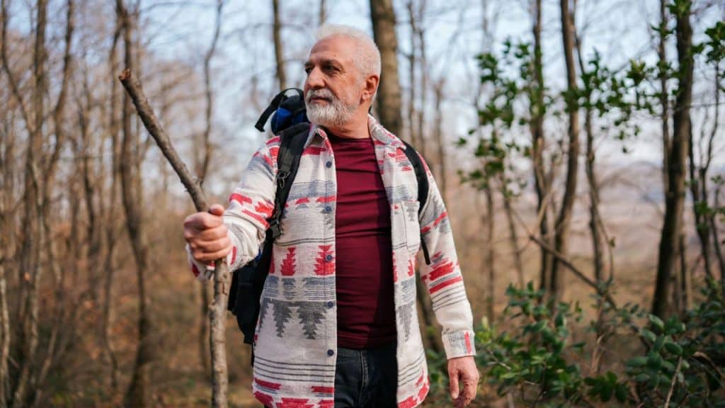 A serious, senior male hiker with a white beard is holding a stick in a forest.