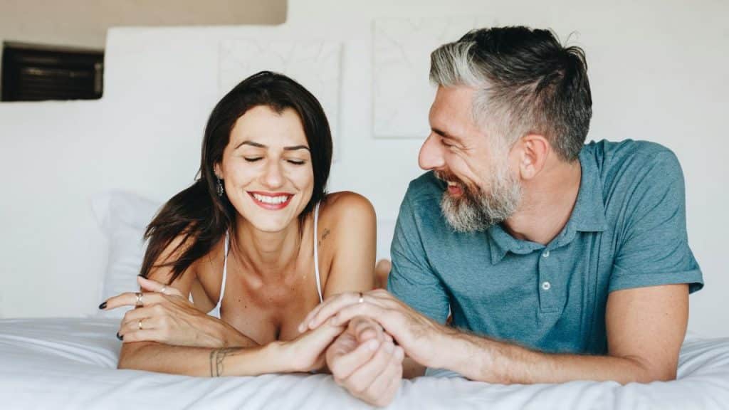 A joyful, mature couple lies on a bed, smiling and holding hands while looking at each other.