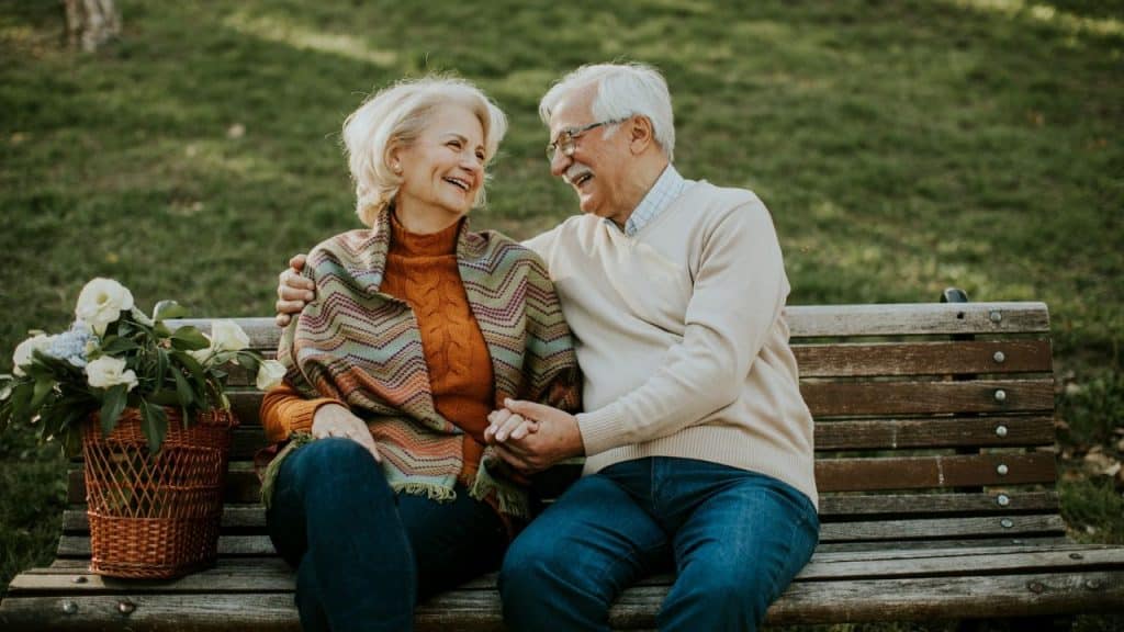 A senior couple is sitting on a bench, smiling at each other and holding hands.