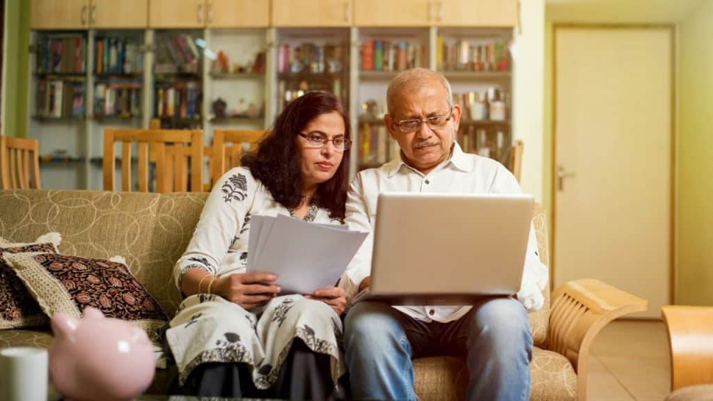 A serious-looking middle-aged couple is sitting on a sofa, looking at a laptop and holding documents.