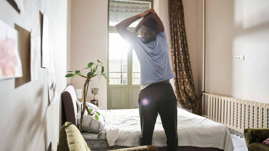 A man stretches his arms over his head while standing next to his bed in a sunlit room.