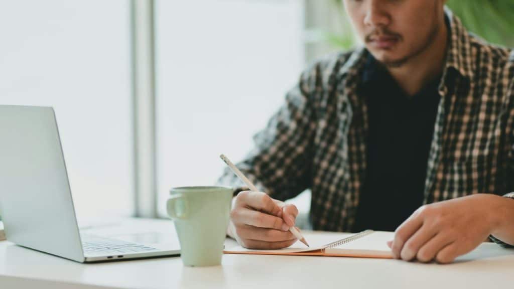 A person with a pencil in their hand is writing in a notebook next to a laptop.