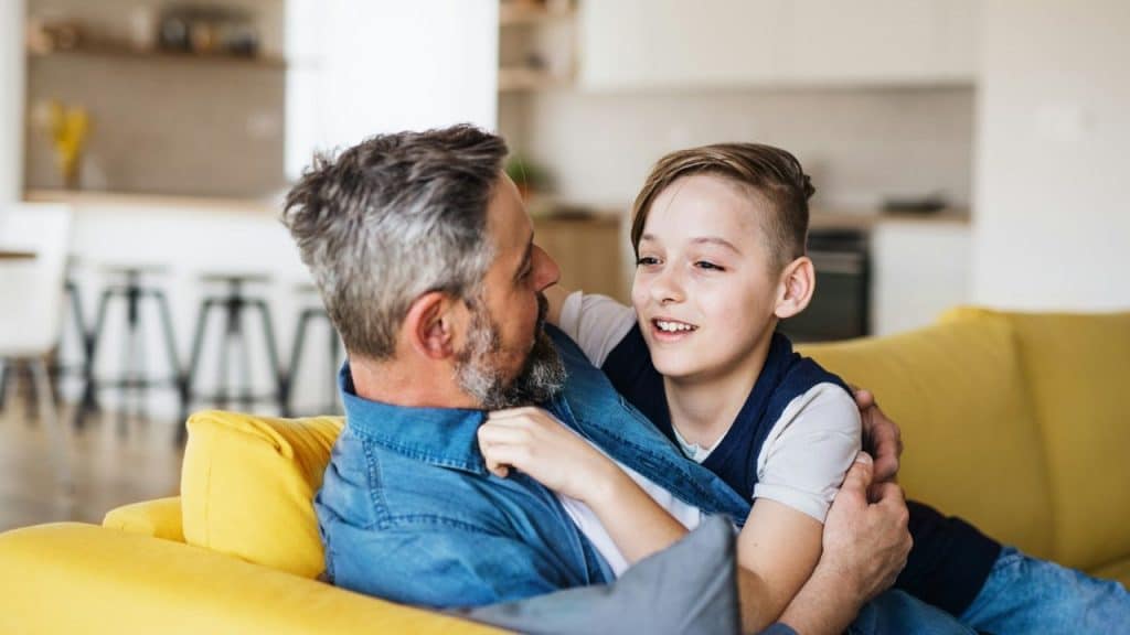 A loving father and his son are sitting on a yellow sofa and talking to each other.