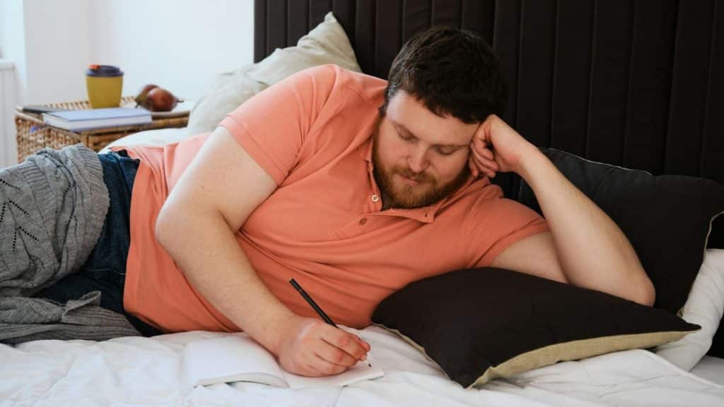 A thoughtful, bearded man is lying on his bed, writing in a journal with a pencil.