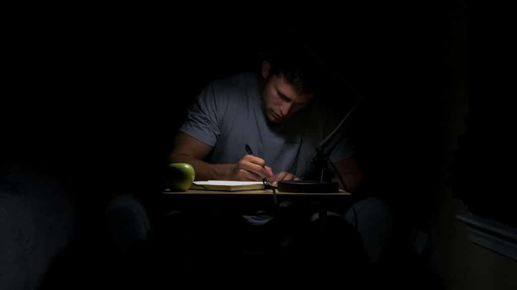 A serious man in a gray T-shirt is writing in a notebook under a desk lamp in a dark room.