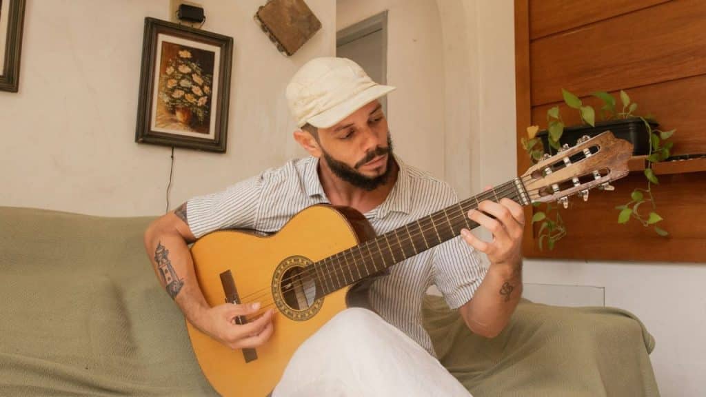 A focused, bearded man wearing a white cap is sitting on a sofa and playing a guitar.