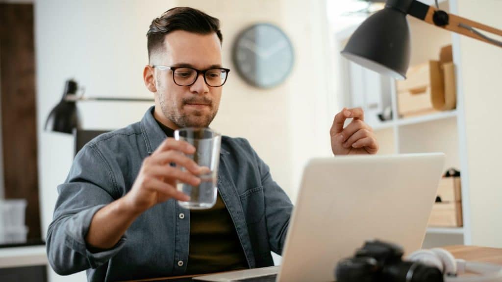 A bespectacled man is sitting at a desk, looking at his laptop and holding a glass of water.