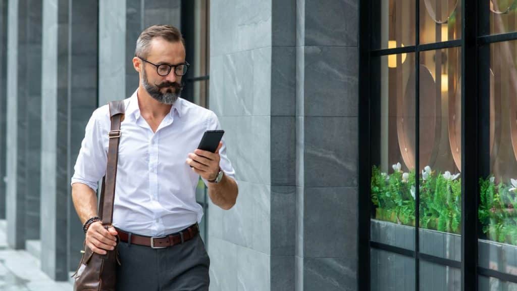 A well-dressed, bearded man with glasses is walking on a city sidewalk and looking at his phone.