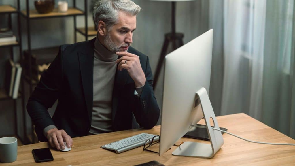 A thoughtful, well-dressed man with a grey beard is working on a computer at a desk.