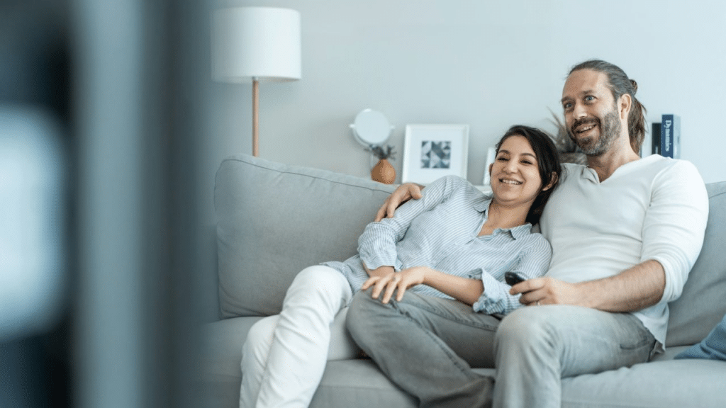 A smiling couple relaxes on a sofa, watching TV and holding a remote.