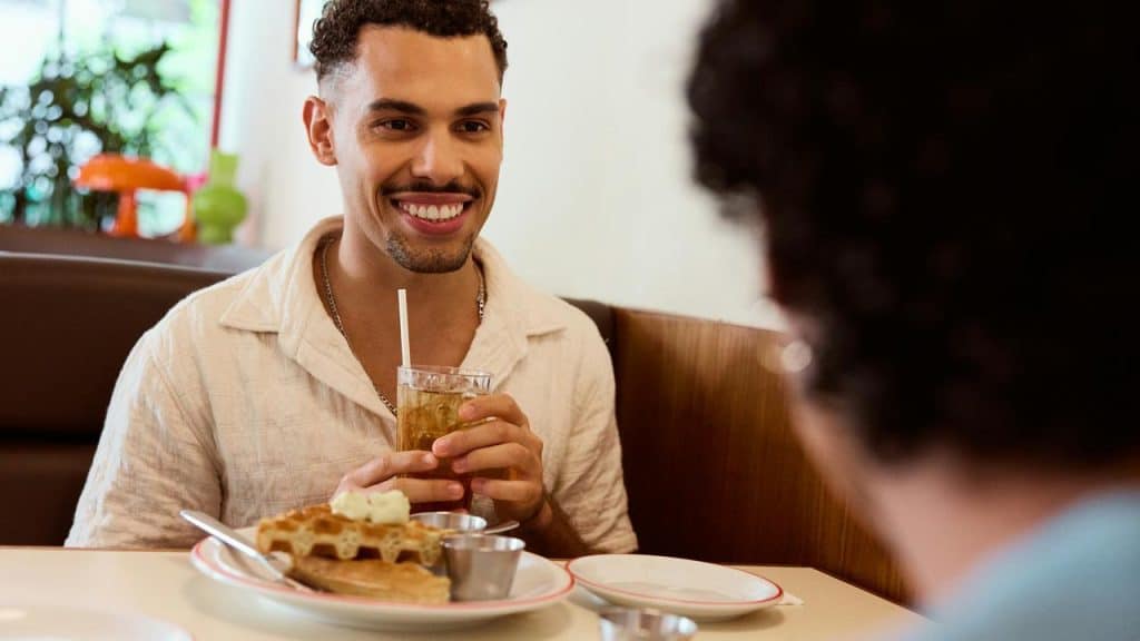 A man smiling and holding a drink while sitting across from someone at a diner.