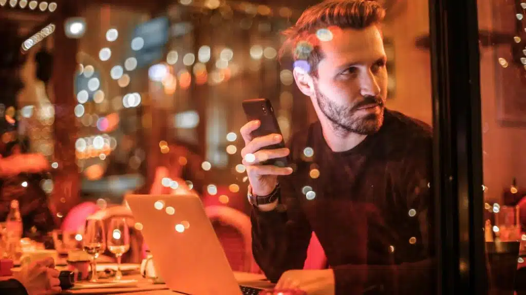 A man holding a phone while sitting at a table with a laptop in a warmly lit café.