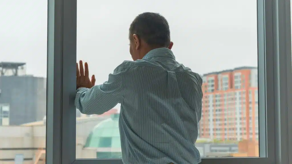 A man in a striped shirt standing by a window with his hand on the glass.