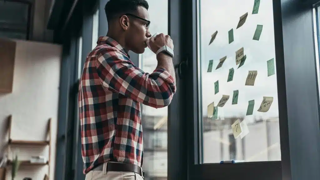 A man drinking from a cup while looking at sticky notes on a window.