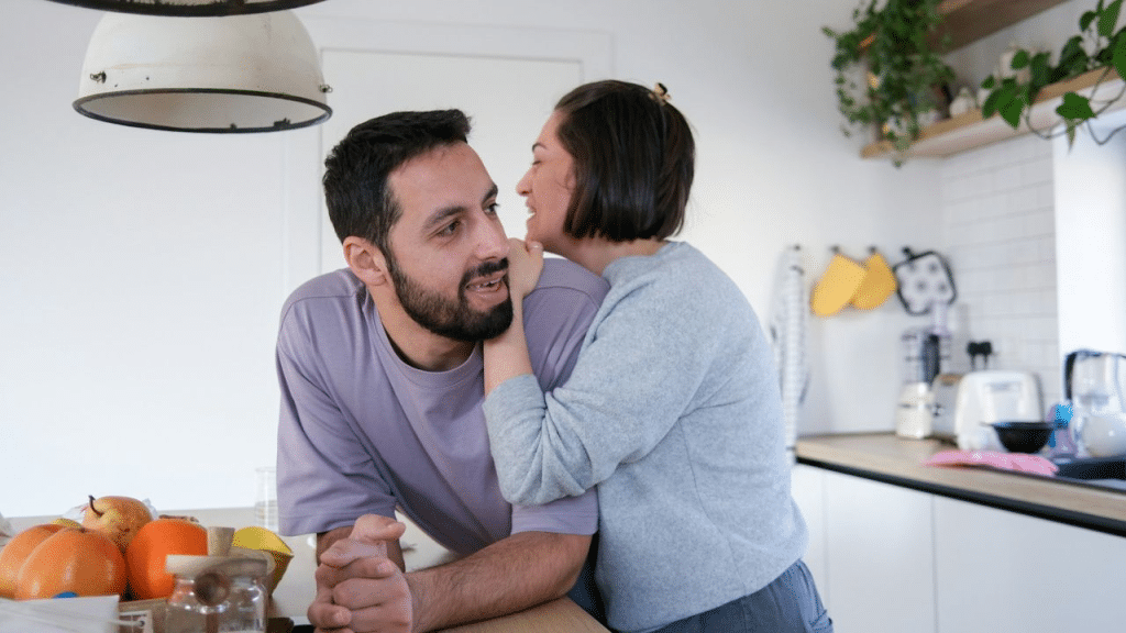 A couple smiles at each other in a kitchen as the woman whispers in the man's ear.