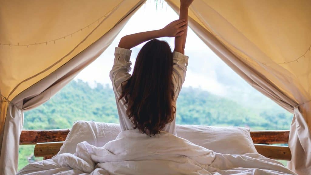 A woman stretching while sitting on a bed inside a tent with a scenic mountain view outside.