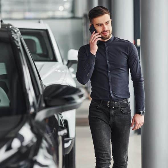A man in dark clothing talks on a phone while walking past cars indoors.