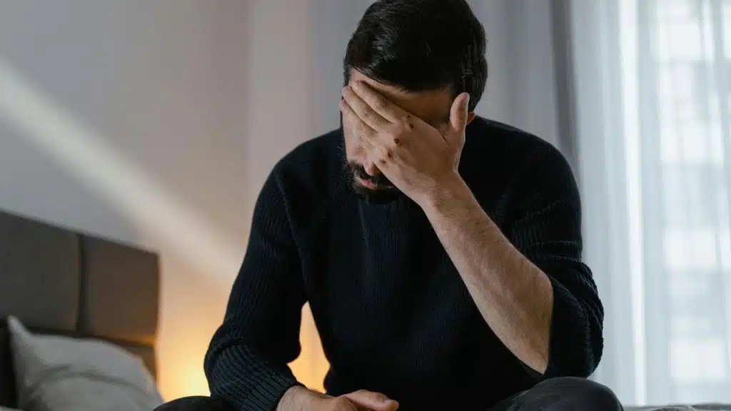 A man sitting on a bed covering his face with his hand in distress.