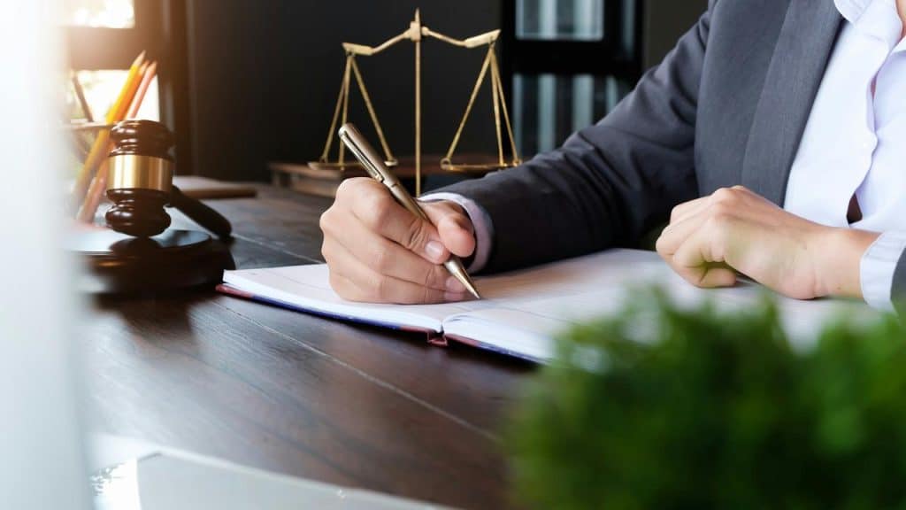 A person in a suit writing in a notebook with legal symbols on the desk.
