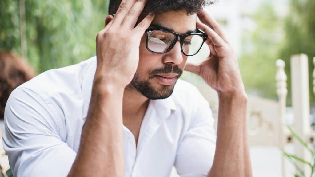A stressed man wearing glasses holding his head with both hands.