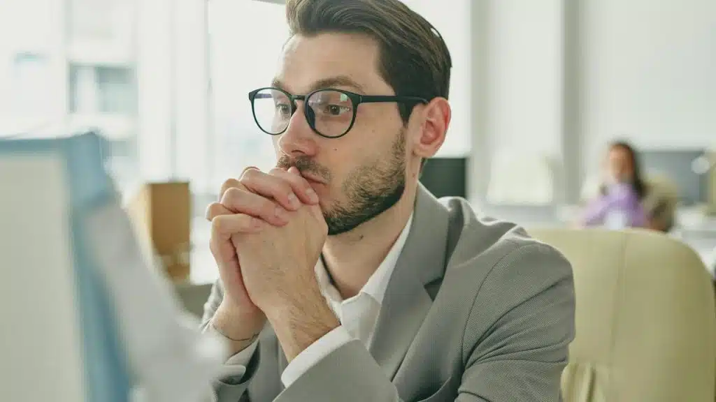 A worried man in glasses and a suit sitting at his desk with hands clasped near his face.