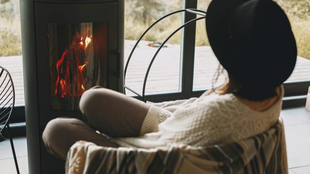 A person sitting by a fireplace wearing a hat and cozy sweater.