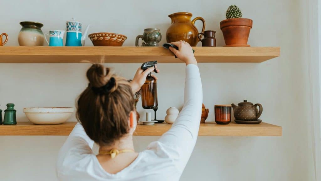 A person cleaning wooden shelves with dishes and pottery.
