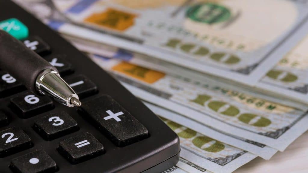 A close-up of a calculator, pen, and stacks of hundred-dollar bills.