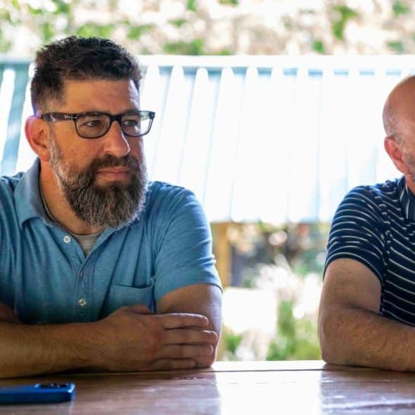 Two men with beards sit at a wooden table outdoors.