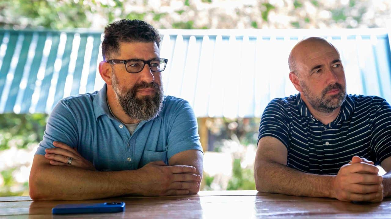 Two men with beards sit at a wooden table outdoors.