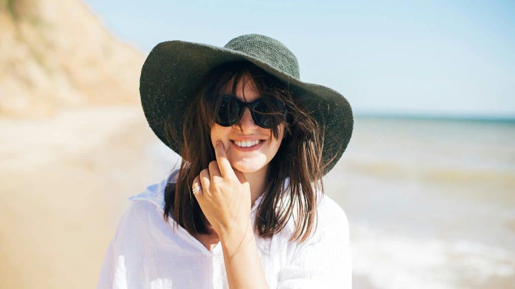 A woman smiling at the beach wearing sunglasses and a wide-brimmed hat.