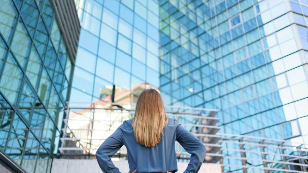 A woman standing with hands on hips looking up at a modern glass building.