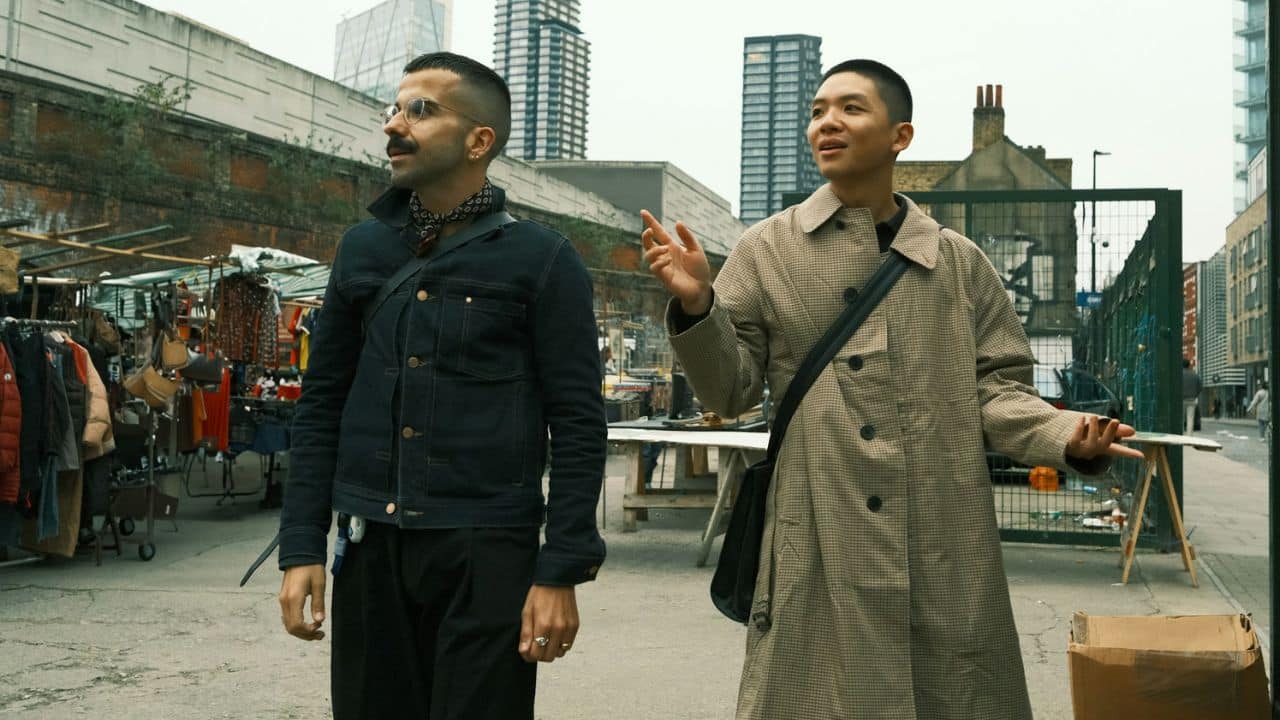 Two men, one in a denim jacket and the other in a plaid trench coat, are walking and talking at a market.
