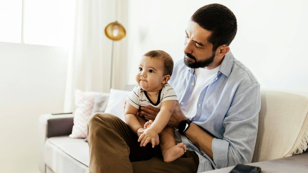 A bearded man sits on a couch, holding an adorable baby on his lap.