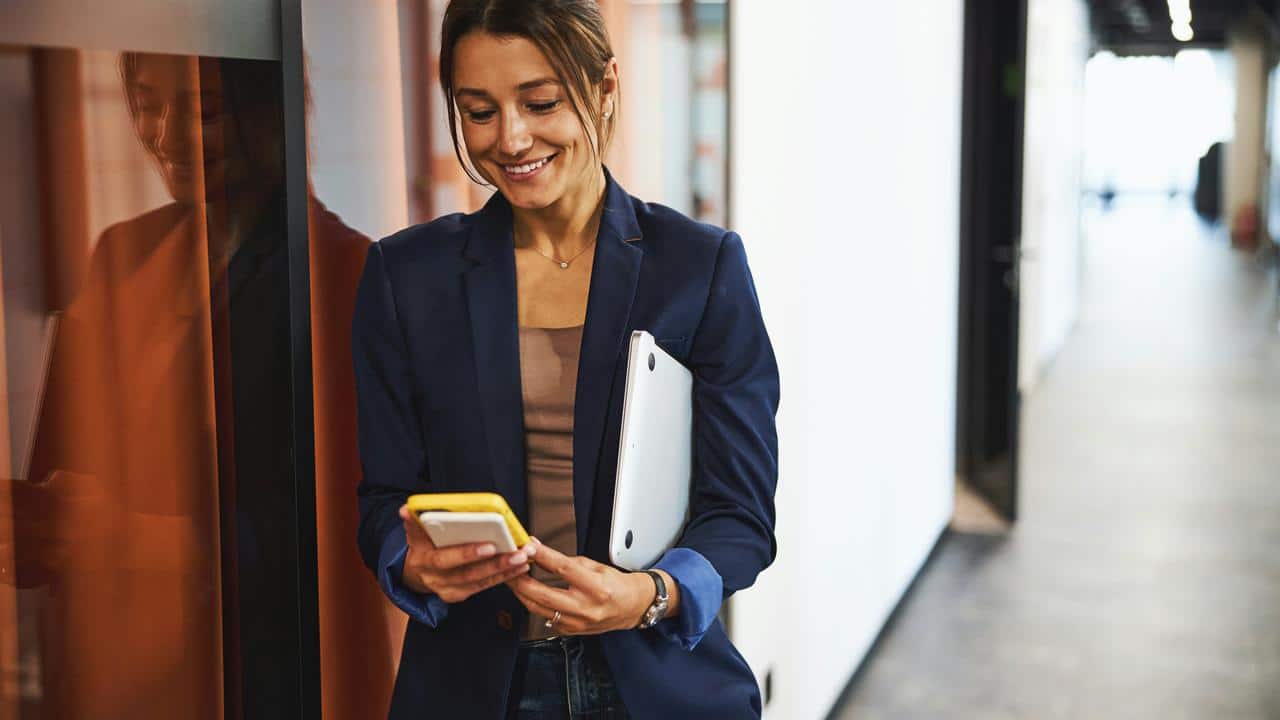 A woman in a blazer smiling at her phone while holding a laptop.