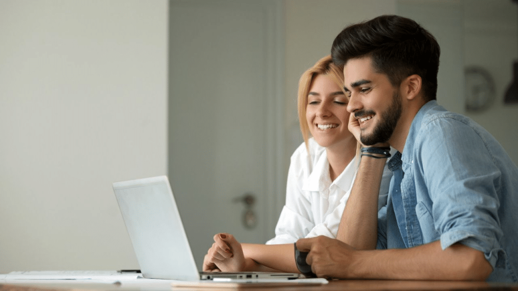A cheerful young couple sits together, looking at a laptop and smiling.