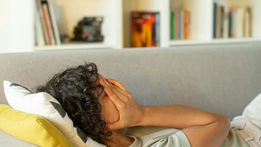 A person lying on a couch covering their face with both hands.
