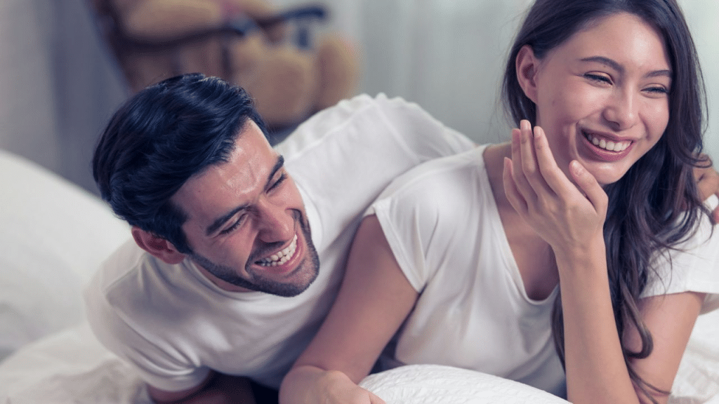 A happy couple in white t-shirts lies on a bed, laughing together.