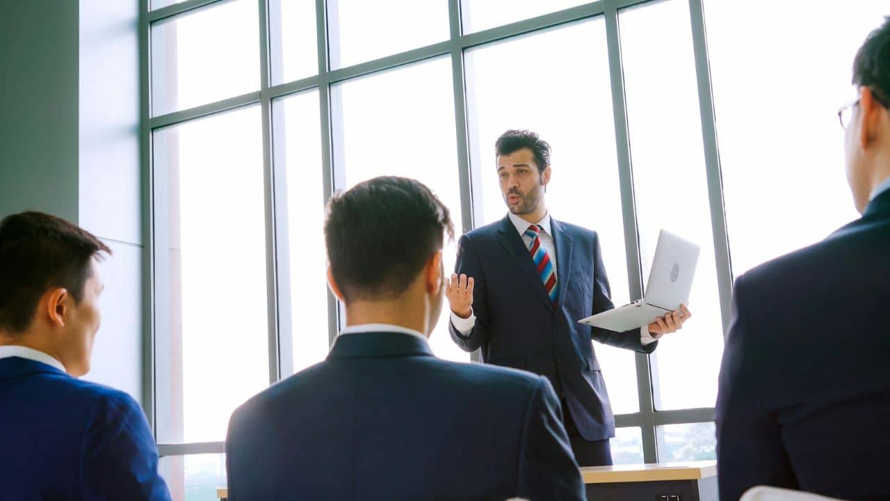 A businessman with a laptop is giving a presentation to three other men in a meeting room.